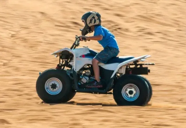 Young boy in a helmet riding a small white kids quad bike on soft sand