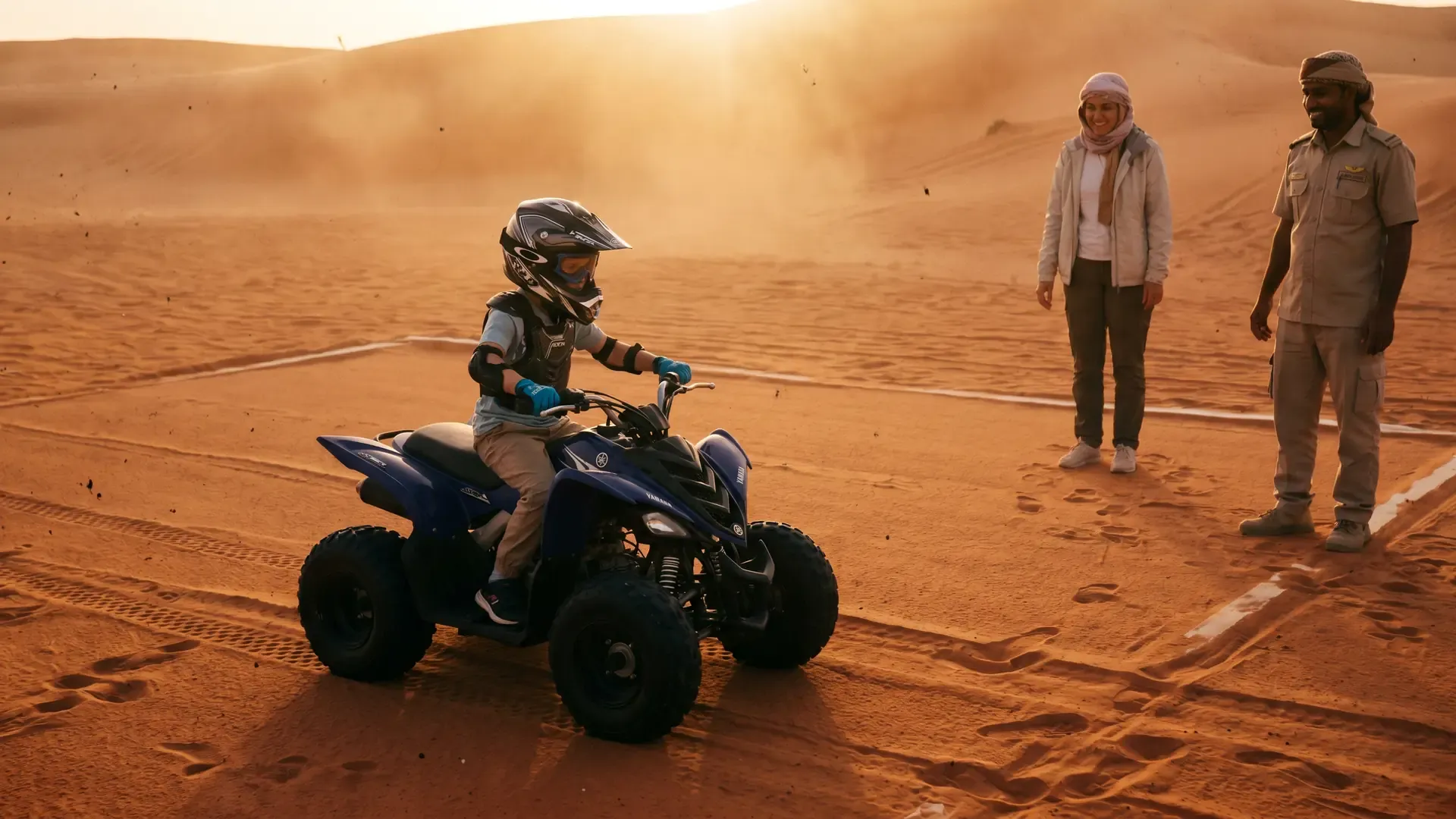 Child in a helmet and gloves on a blue kids quad bike while an adult watches from the sand
